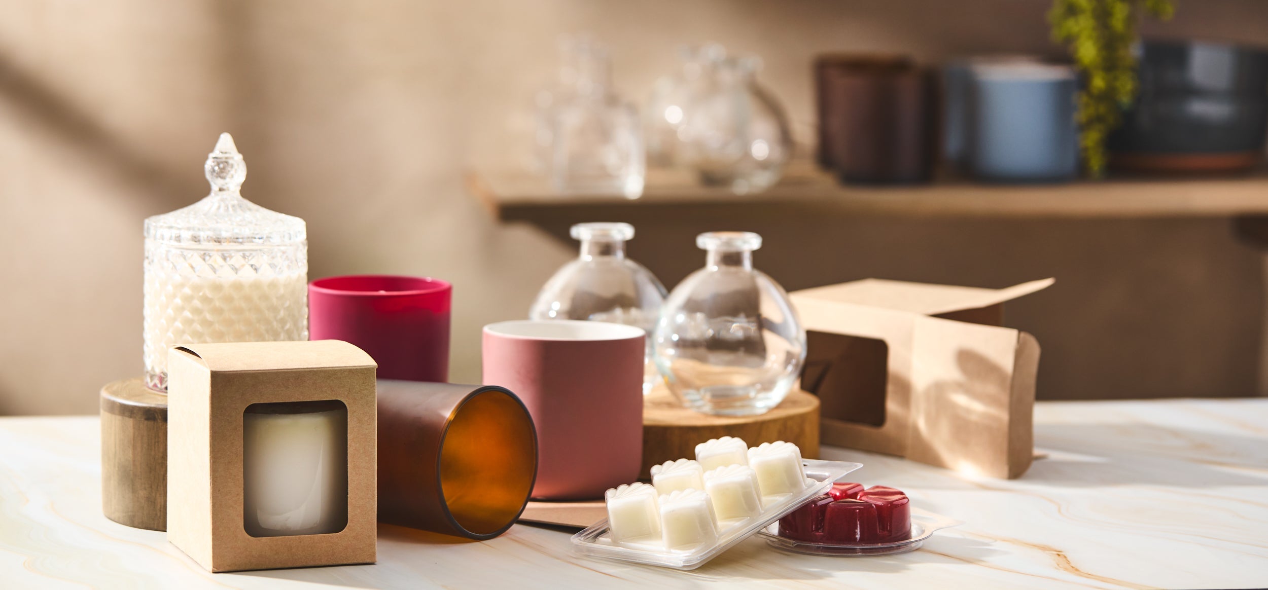 Candle jars, wax melts, and kraft boxes arranged on a marble surface with warm light. A mix of glass, frosted, and colored containers sits beside packaging supplies in a cozy, creative workspace.