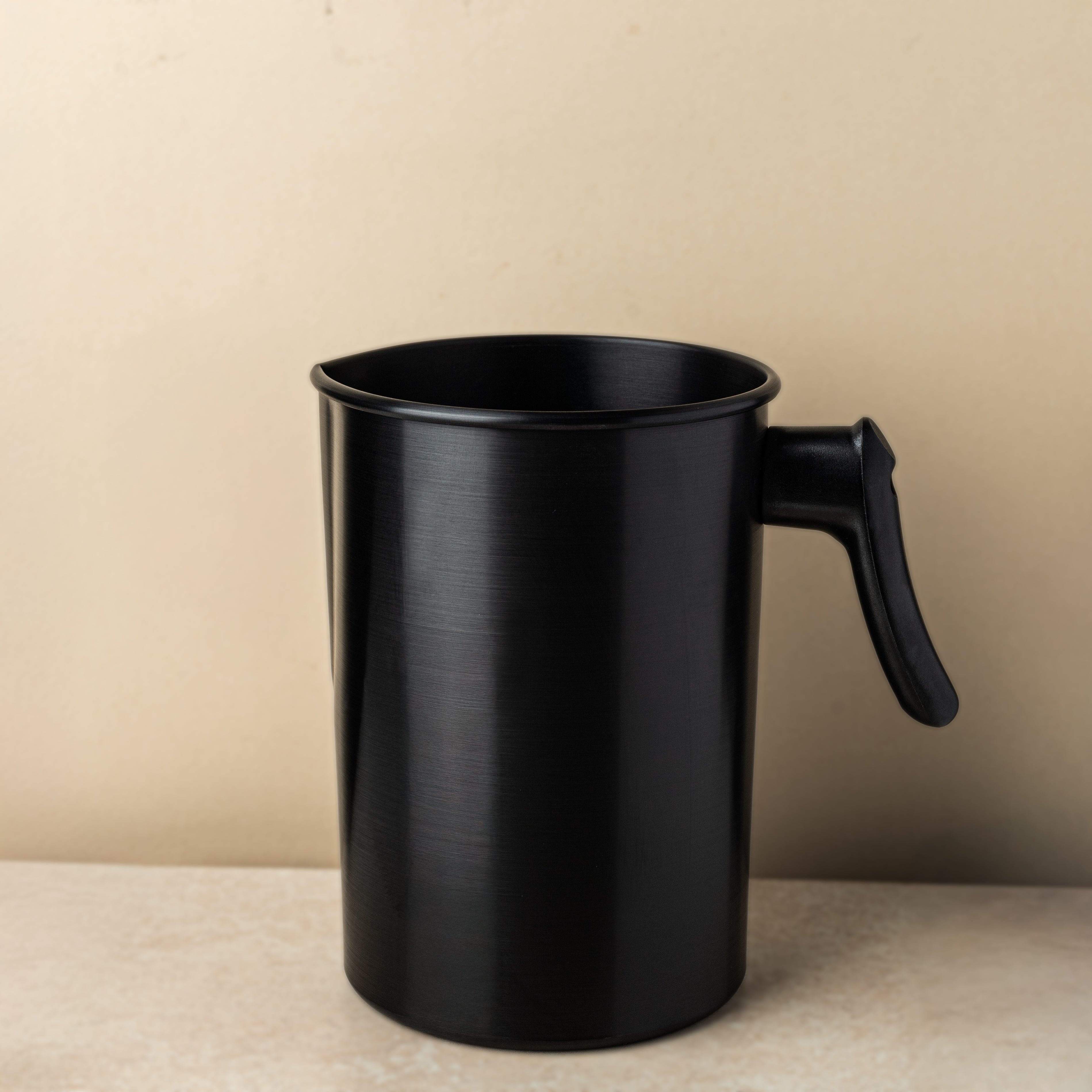 Black metal pouring pitcher with a handle, used for candle making, set against a neutral beige background on a light stone surface.