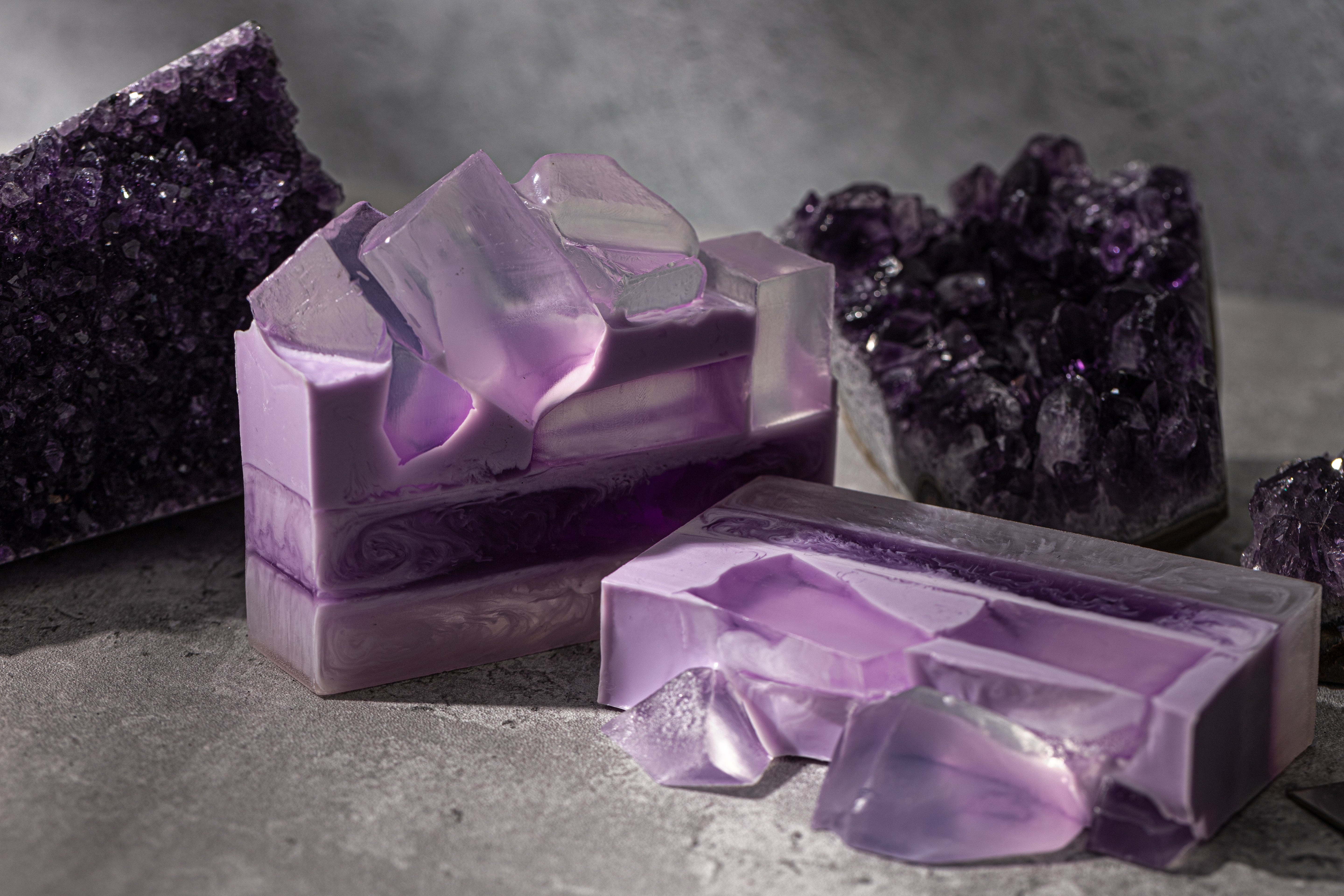 Purple gemstone-inspired handmade soaps with translucent layers and crystal-like tops, styled alongside amethyst geodes on a textured gray surface.