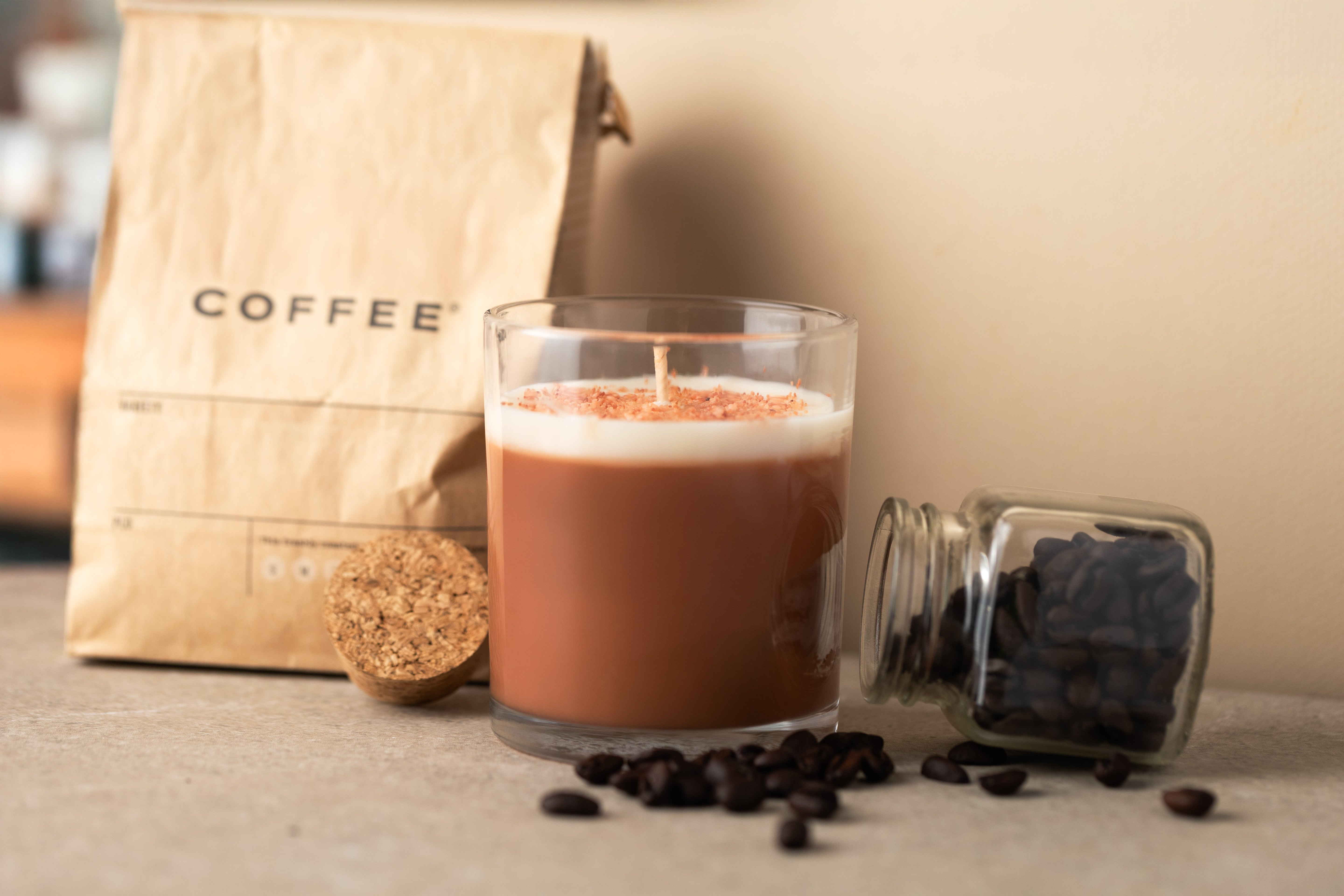 Coffee-scented candle in a glass jar surrounded by coffee beans, a cork lid, and a brown paper coffee bag, with an open glass jar of beans tipped nearby on a neutral background.