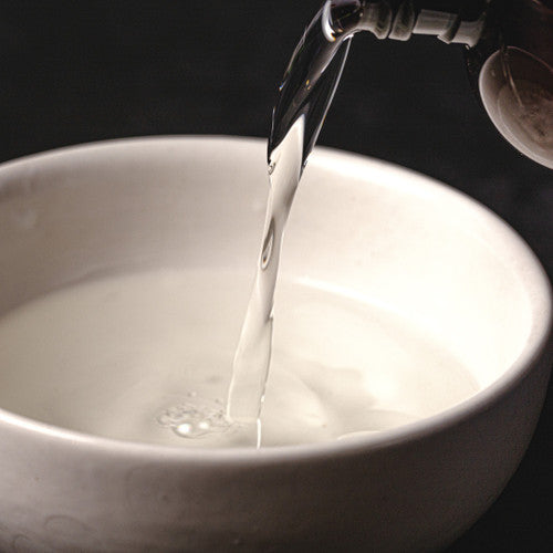 Clear liquid body spray base being poured into a white ceramic bowl, with a smooth stream captured mid-pour against a dark background.