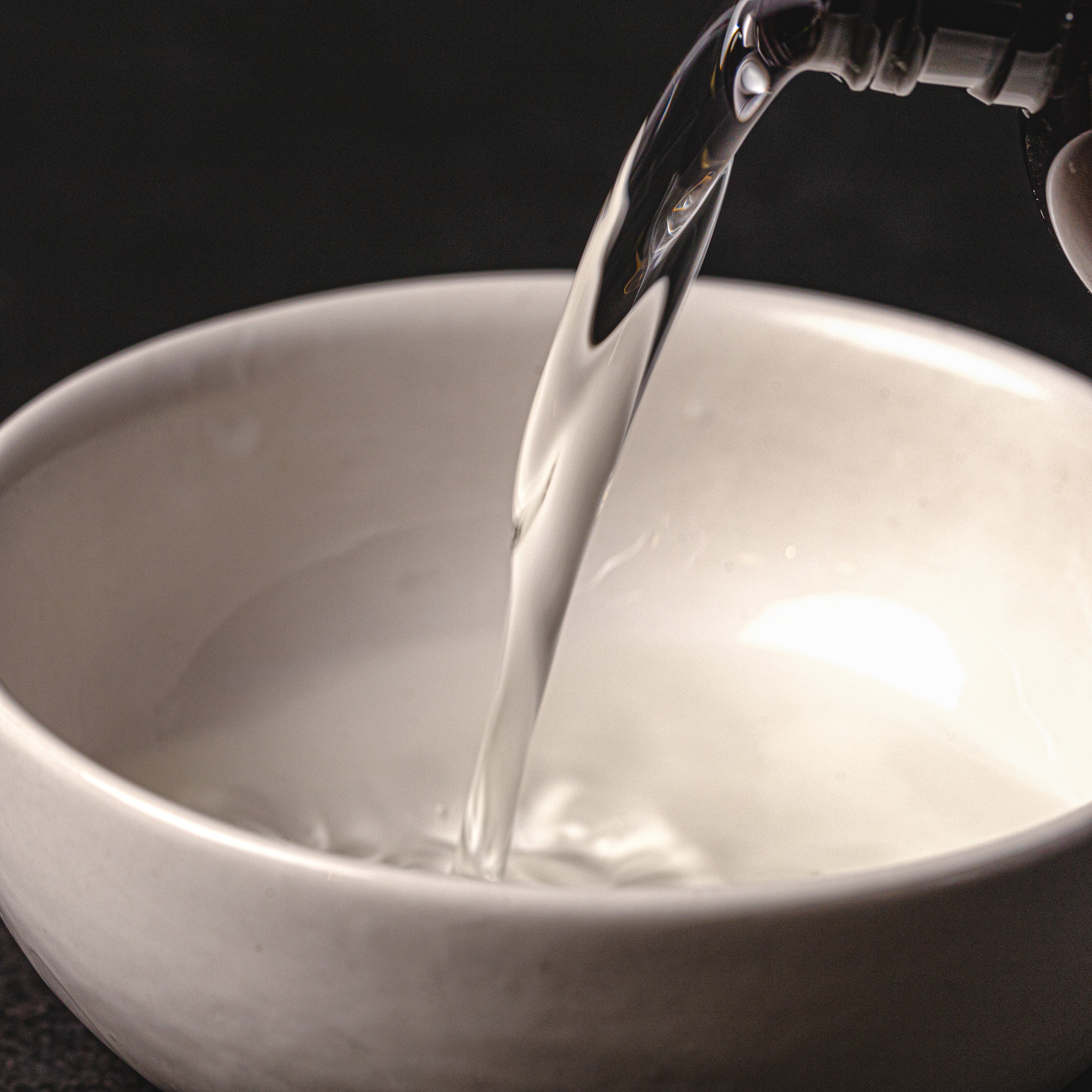Clear liquid body spray base being poured into a white ceramic bowl, with a smooth stream captured mid-pour against a dark background.