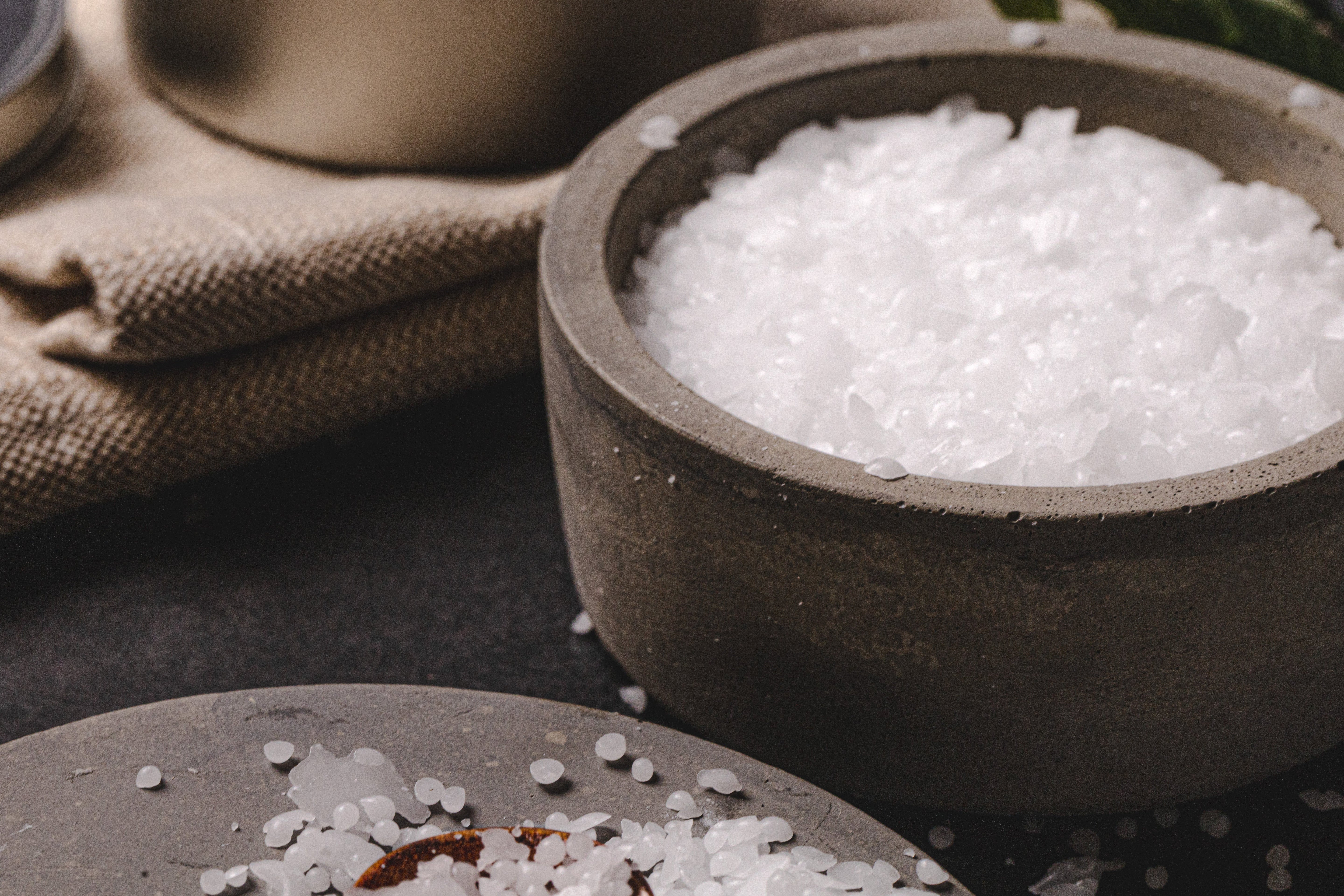 Close-up of white paraffin wax flakes in a gray concrete bowl on a dark surface with a linen cloth and candle-making tools in the background.