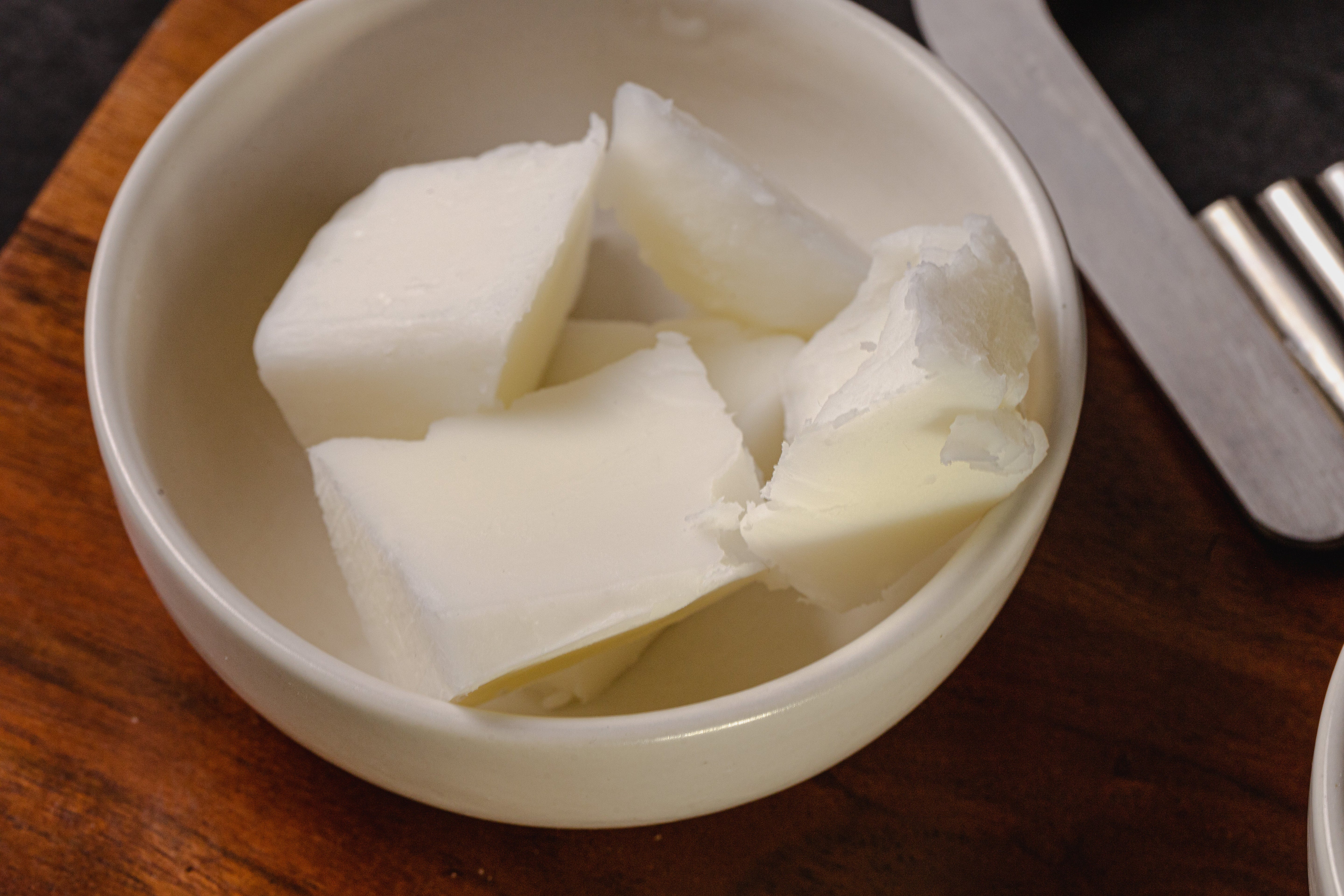 Close-up of creamy white wax blend chunks in a small white ceramic bowl on a wooden surface, with a metal tool partially visible nearby.