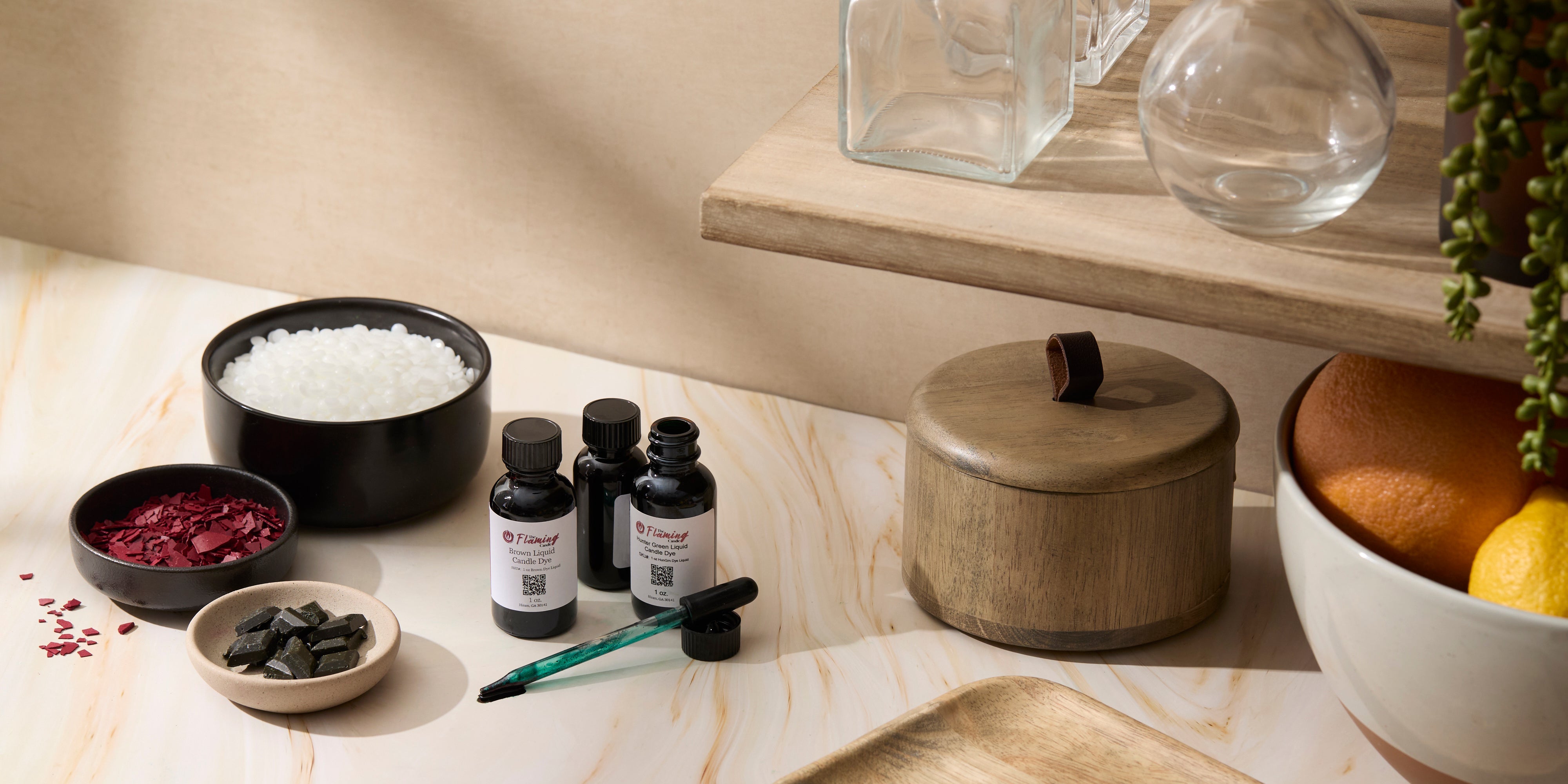 A bright workspace with candle-making additives including bottles of liquid dye, bowls of wax beads and colored flakes, and small wax pieces. The scene features natural light, wood accents, and glass jars for a clean, crafted look.