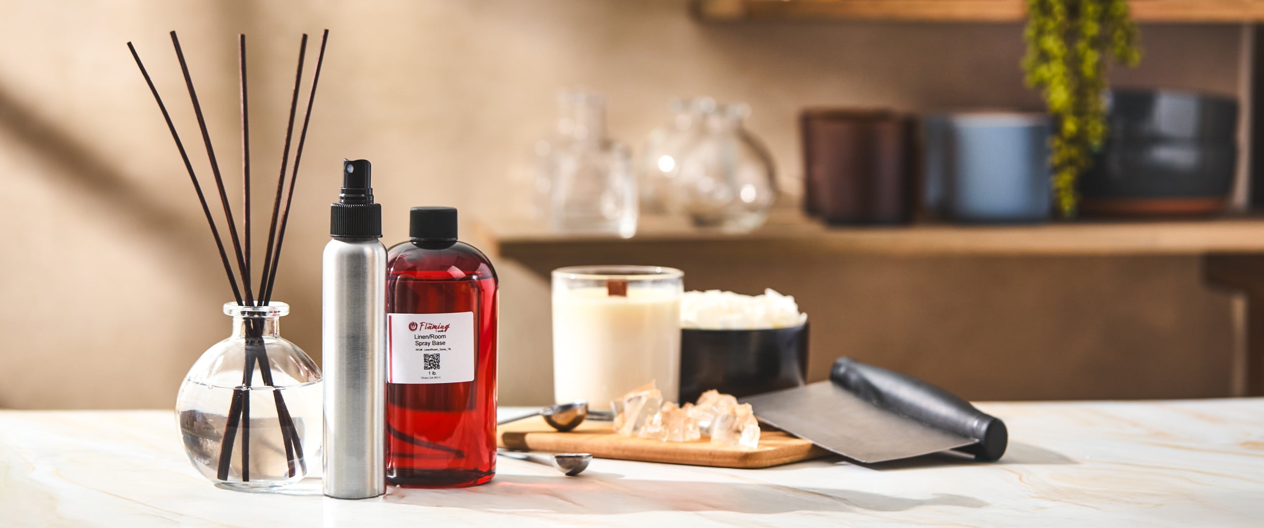A clean, sunlit workspace featuring home fragrance supplies including a reed diffuser, spray bottle, and red bottle labeled “Linen/Room Spray Base,” with a candle, wax pieces, and tools arranged neatly on a marble surface.