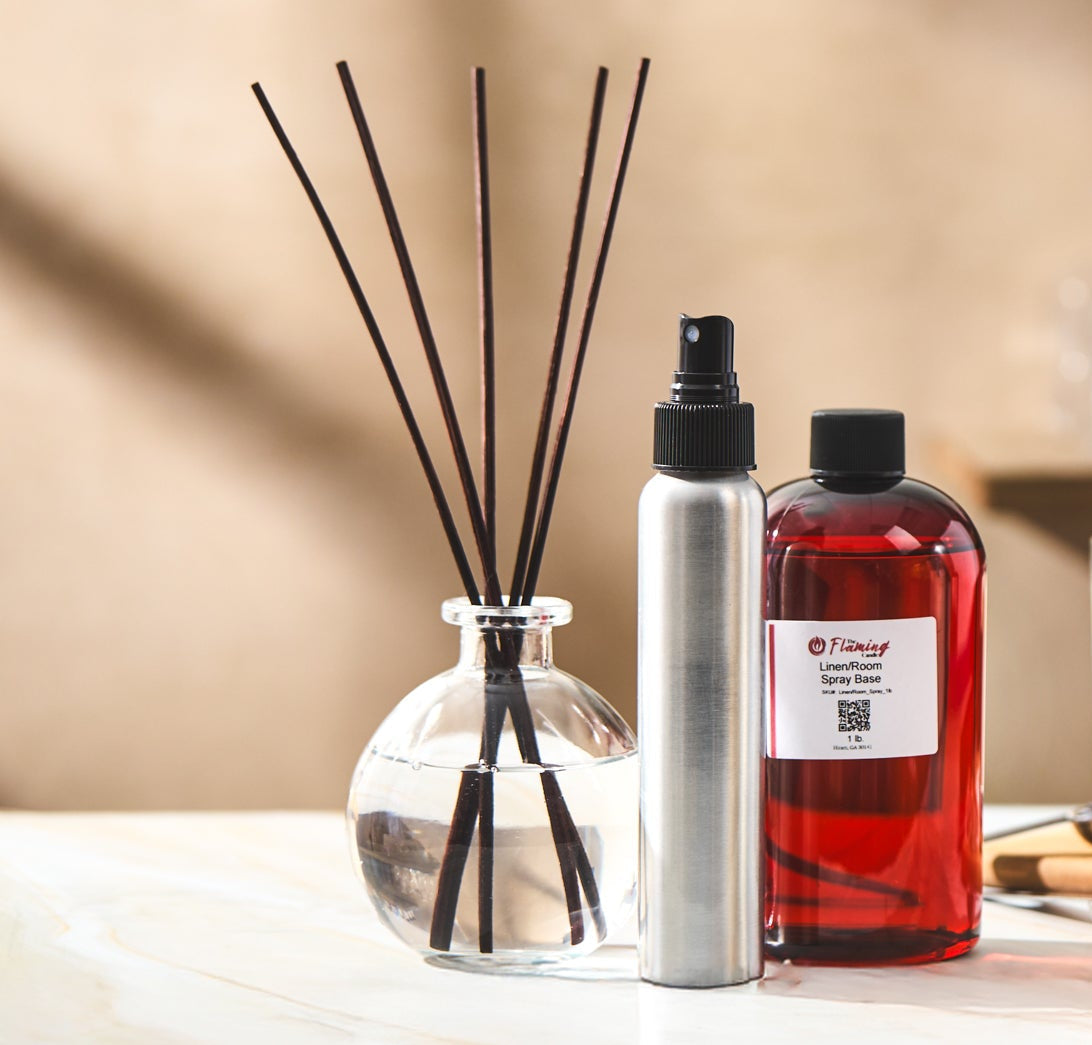 Home fragrance setup featuring The Flaming Candle “Lemon Blossom” fragrance oil, a sleek silver spray bottle, and a clear glass reed diffuser with black reeds on a marble surface against a soft beige background.