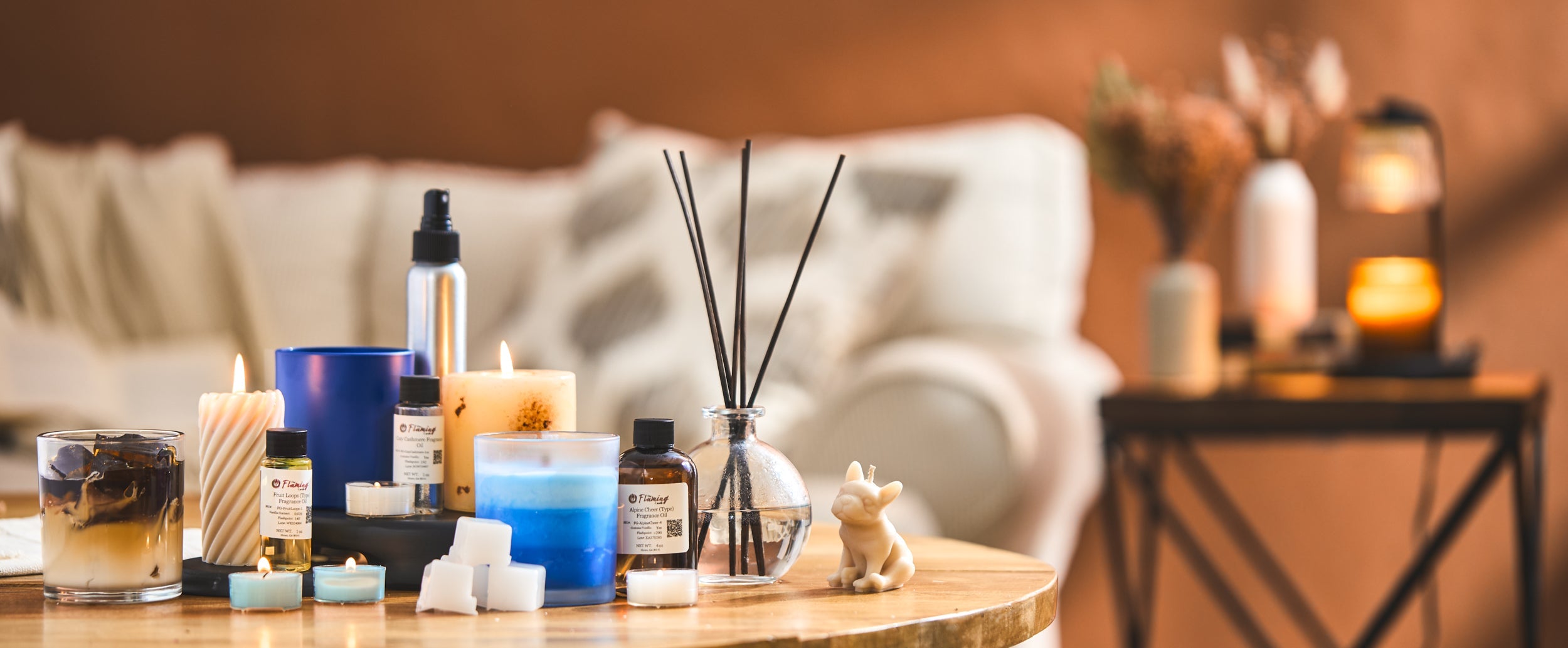 Candles, fragrance oils, wax melts, and reed diffusers displayed on a wooden table in a cozy living room. A soft-focus background with a couch and glowing candles creates a warm, inviting atmosphere.