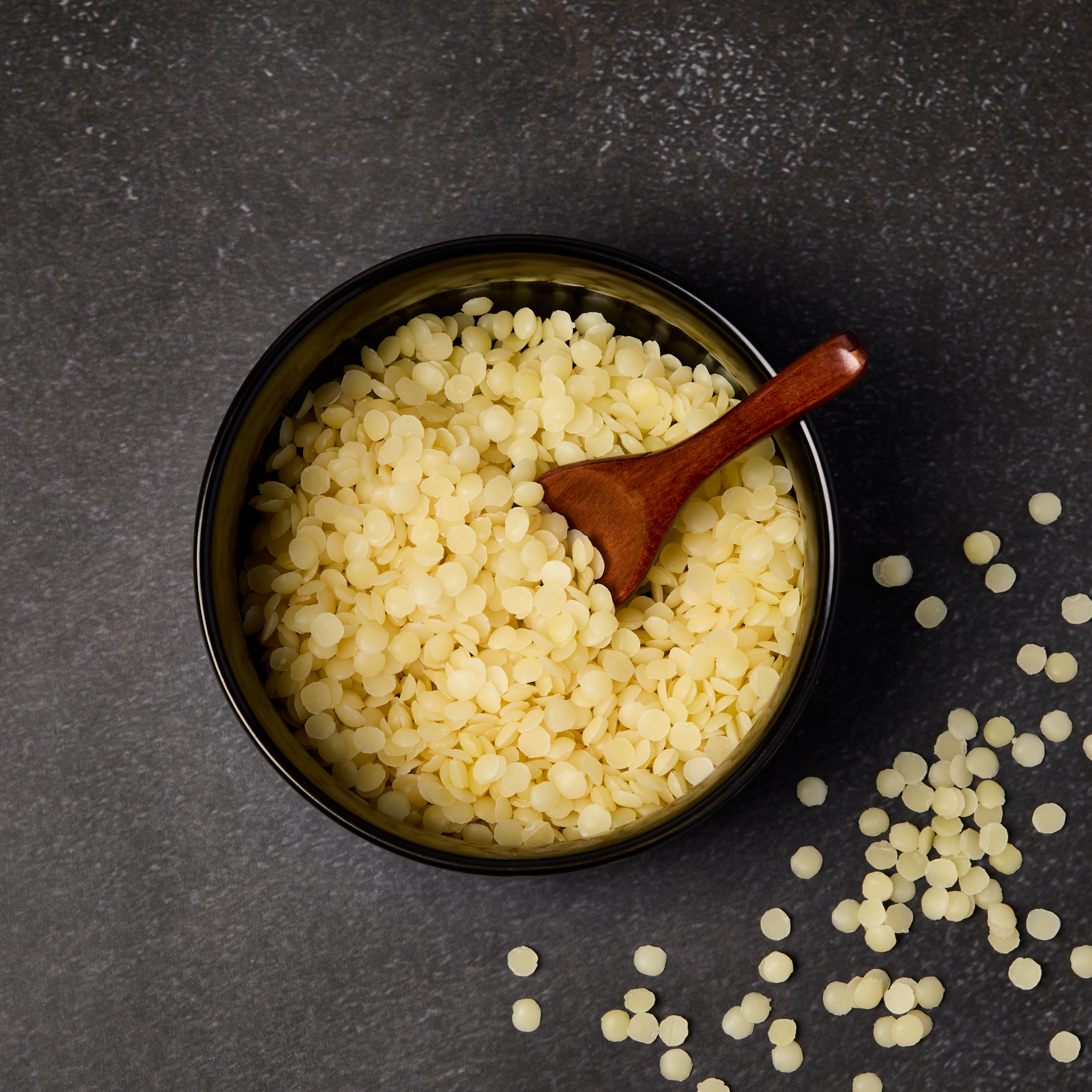 Overhead view of natural yellow beeswax pellets in a black bowl with a small wooden spoon on a dark textured surface, with a few pellets scattered around.
