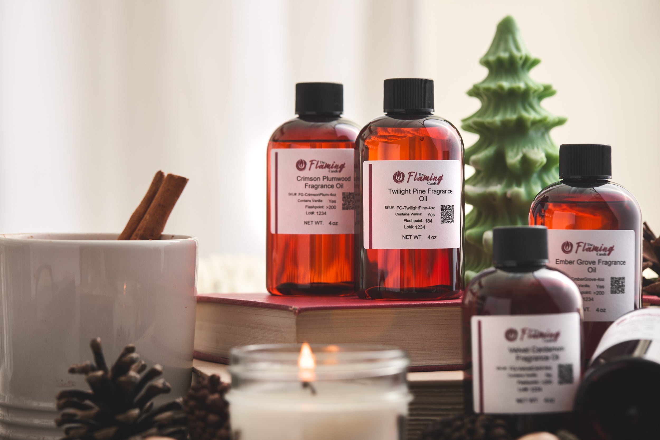Alt text: Cozy holiday setup featuring The Flaming Candle fragrance oils labeled “Crimson Plumwood,” “Twilight Pine,” and “Ember Grove,” surrounded by pinecones, candles, books, and a green tree-shaped candle.