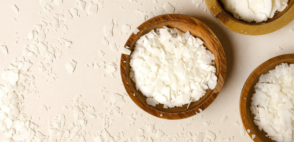 Wooden bowls filled with white soy wax flakes are arranged on a light surface, with scattered flakes adding texture and a natural, handcrafted feel.