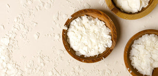Wooden bowls filled with white soy wax flakes are arranged on a light surface, with scattered flakes adding texture and a natural, handcrafted feel.
