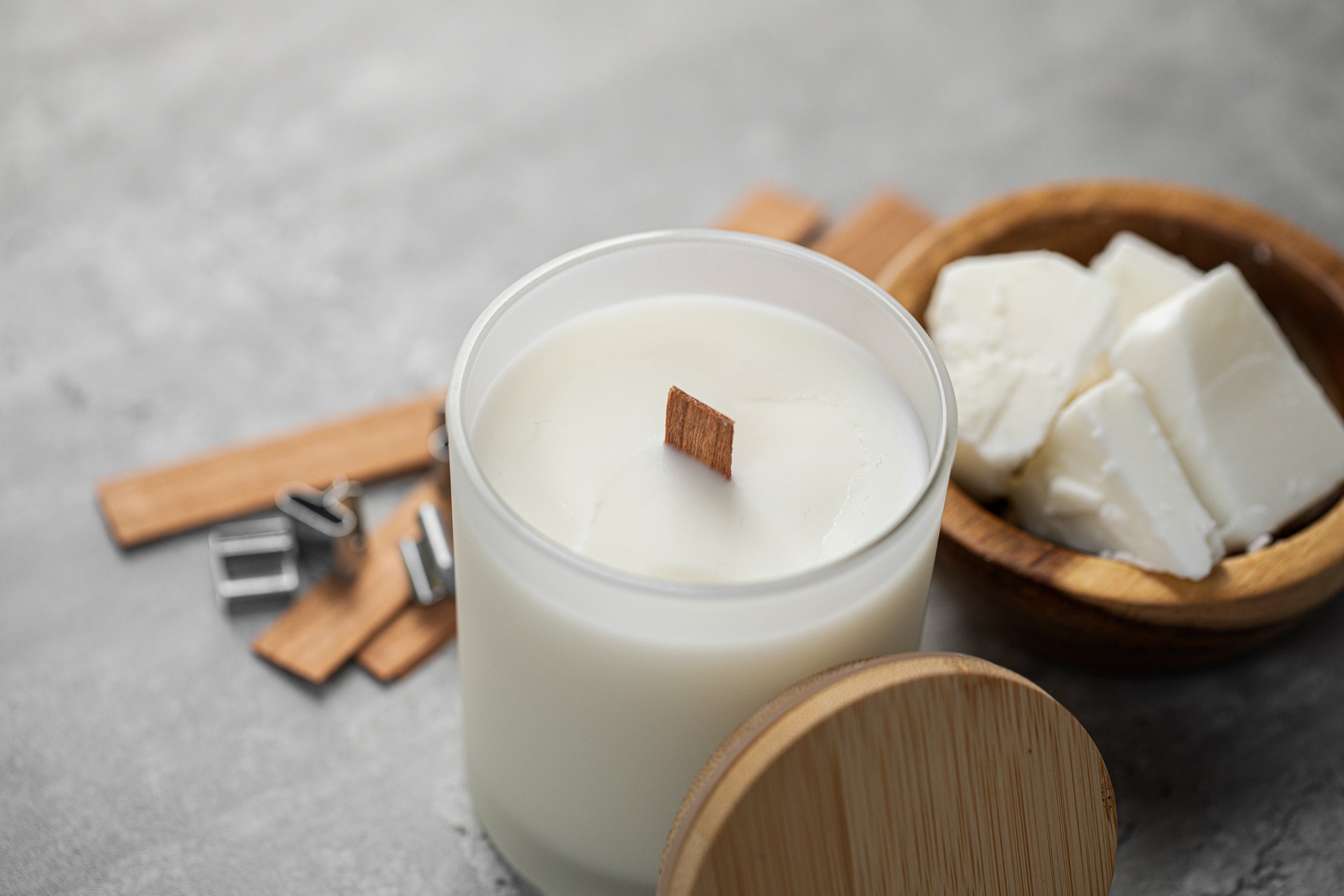 Close-up of a white candle with a wooden wick in a frosted glass jar beside a wooden bowl filled with wax chunks, wooden wick clips, and a bamboo lid on a gray surface.
