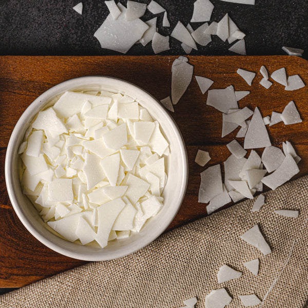 White wax flakes displayed in a ceramic bowl and wooden spoon on a wooden board, with additional flakes scattered around — a clean, natural setup ideal for showcasing candle-making ingredients.