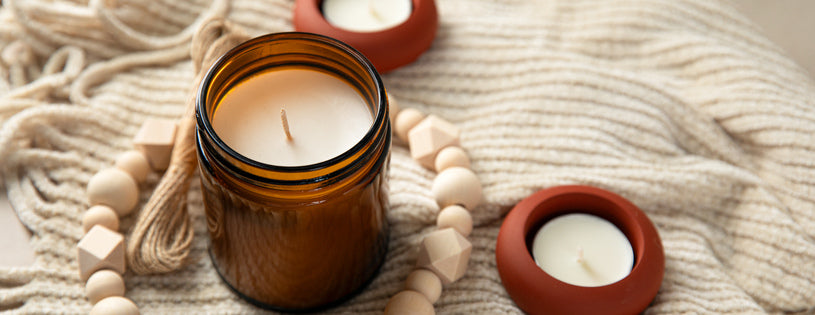 A cozy setup featuring a candle in an amber glass jar surrounded by two small tealights in terracotta holders, wooden beads, and a soft beige knit blanket.