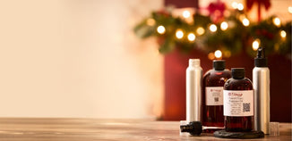 Christmas room spray kit on a wooden table—amber fragrance oil bottles and silver spray bottles in front of soft, warm holiday lights and greenery, with empty space on the left.