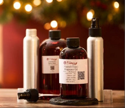 A festive room spray kit featuring The Flaming Candle fragrance oils and aluminum spray bottles, styled on a wooden surface with soft holiday lights and greenery in the background.
