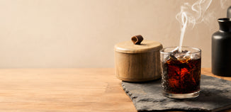 Glass of dark iced beverage with wisps of smoke on a slate board beside a rustic wooden container and black vase.