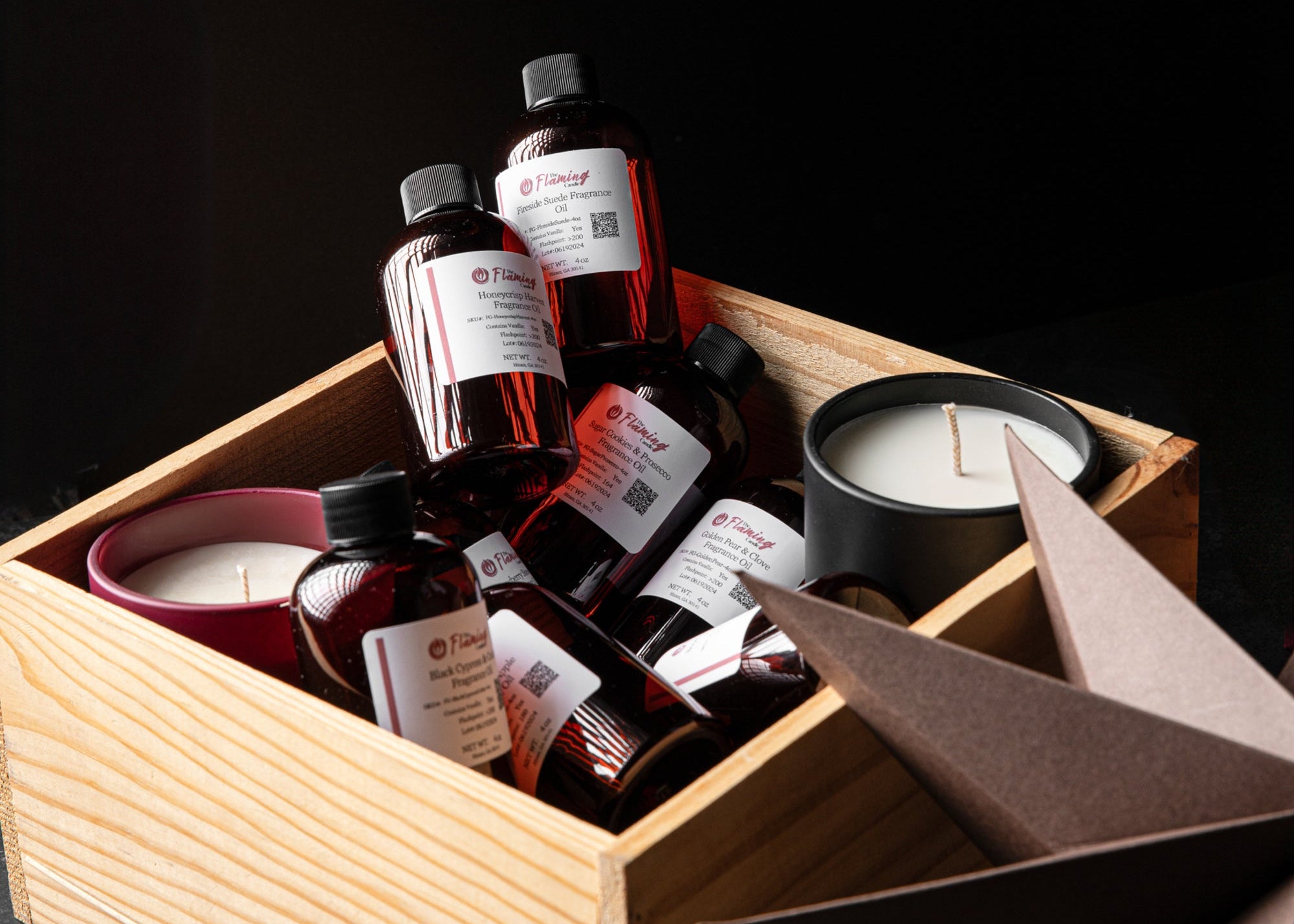 Wooden crate filled with amber fragrance oil bottles from The Flaming Candle and finished candles, styled on a dark background with folded paper star