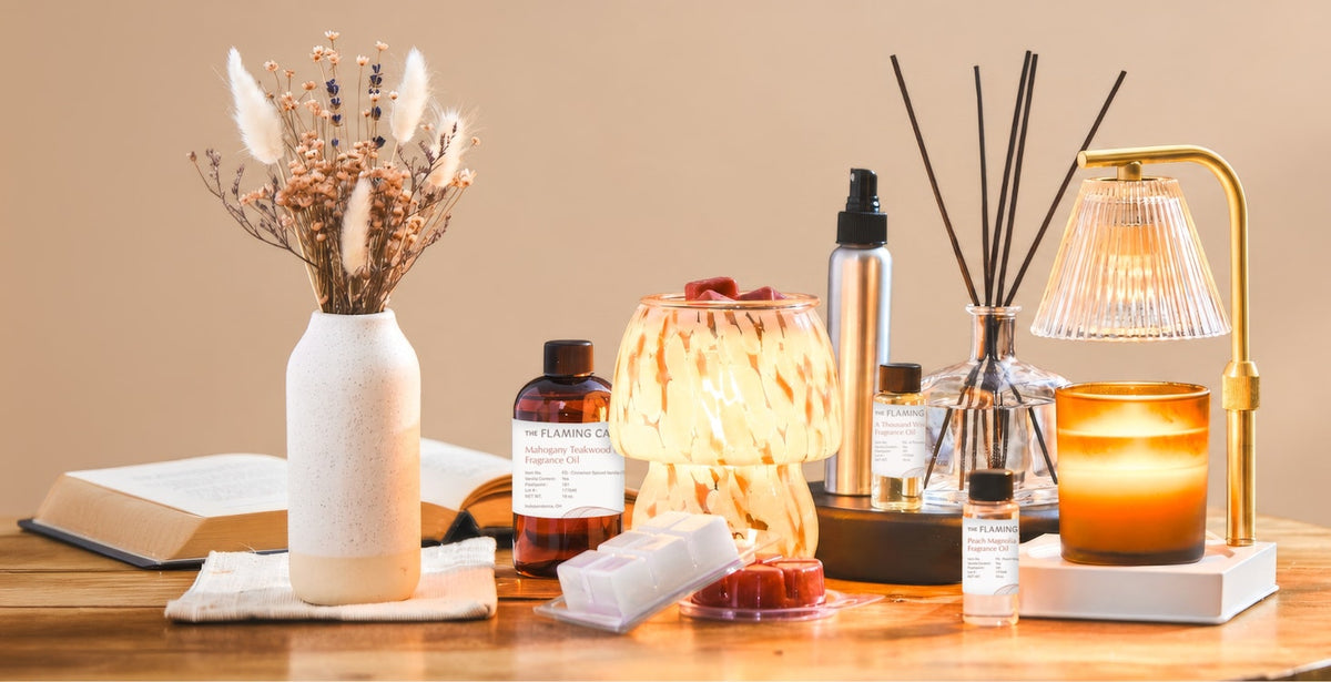Wooden table styled with dried flowers, an open book, glowing candle warmer, fragrance oils, wax melts, reed diffuser, amber candle, and gold lamp against a warm beige background.