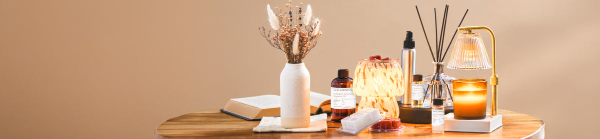 Wooden table styled with dried flowers, an open book, glowing candle warmer, fragrance oils, wax melts, reed diffuser, amber candle, and gold lamp against a warm beige background.