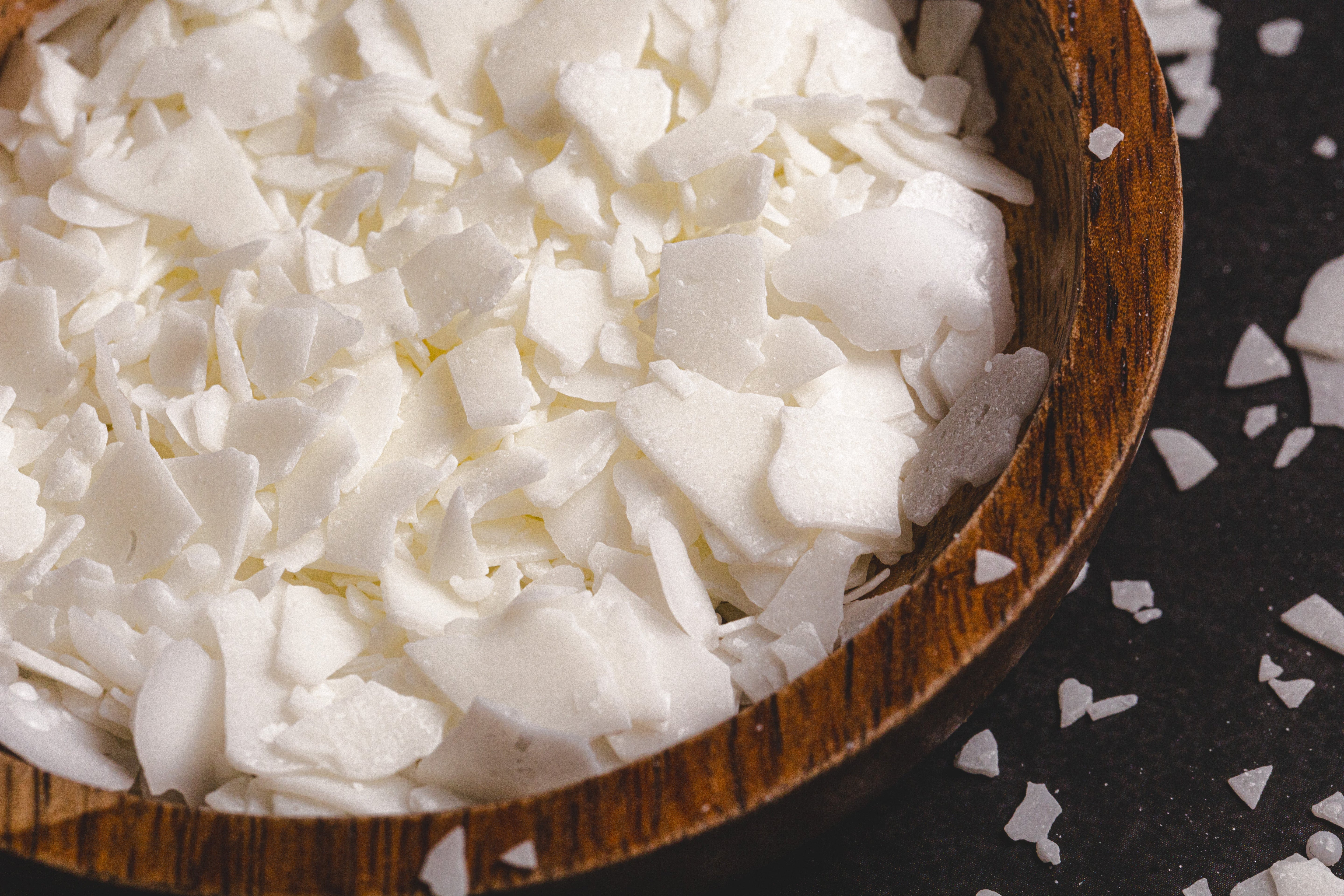 Close-up of natural soy wax flakes in a wooden bowl, with scattered white pieces on a dark textured surface.