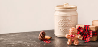 Gingerbread man wax melt with red bow tie and buttons beside a cream cookie jar, plaid ribbon wrapped gifts, pinecone, and cinnamon stick.