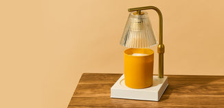 A yellow candle sits beneath a modern glass and brass candle warmer lamp on a wooden surface, creating a cozy, elegant home fragrance display.