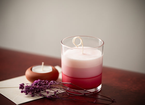 Glass jar candle with pink-to-red gradient wax on a wooden table, styled with a tea light and dried purple flowers.