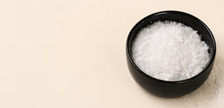 Black bowl filled with white soy wax flakes on a light neutral background, with a small spill of wax flakes beside the bowl and ample copy space.