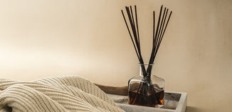 A glass reed diffuser sits beside a soft knit blanket, creating a cozy and inviting home fragrance scene.
