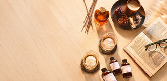 Flat lay of a cozy home fragrance scene with an amber candle, reed diffuser, fragrance oils, whiskey glasses, pinecones, and an open book on a light wood tabletop.