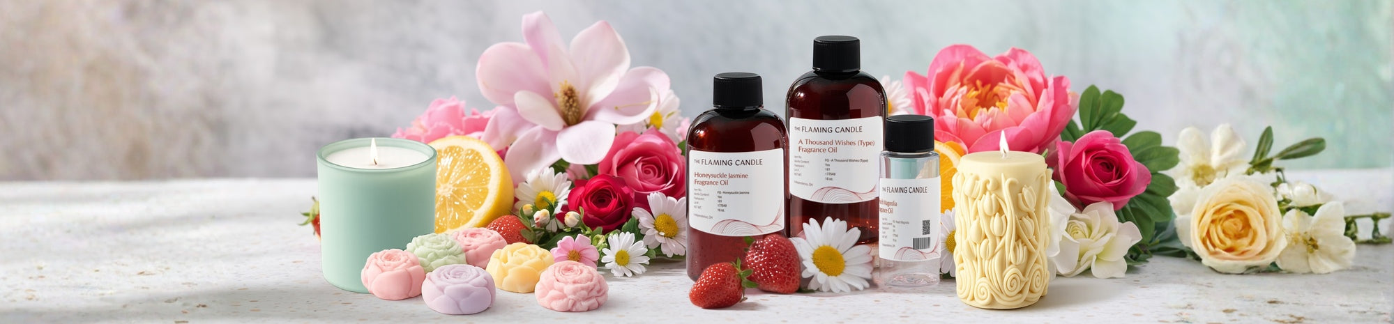 Spring-themed display featuring The Flaming Candle fragrance oils, pastel candles, and wax melts surrounded by fresh flowers, strawberries, and a lemon slice on a light stone surface, creating a bright, floral-inspired scent story.