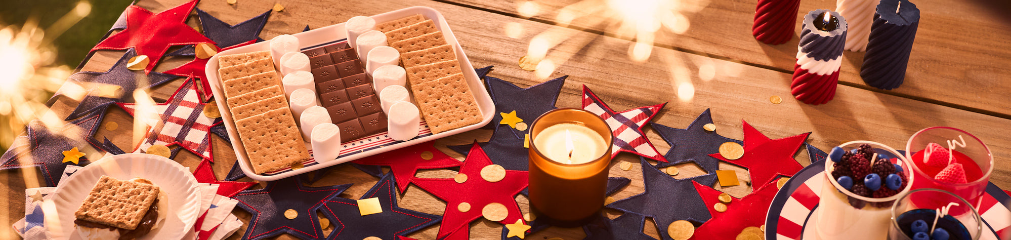 Patriotic dessert spread with graham crackers, marshmallows, and chocolate arranged on a tray, surrounded by red, white, and blue decorations, candles, and sparklers on a wooden table.