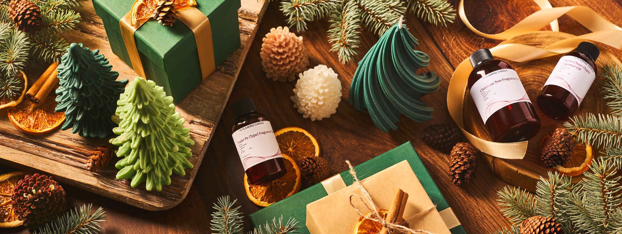 Holiday flat lay with evergreen branches, pinecones, dried orange slices, cinnamon sticks, green gift boxes, tree-shaped candles, and fragrance oil bottles arranged on a rustic wooden table with gold ribbon accents.