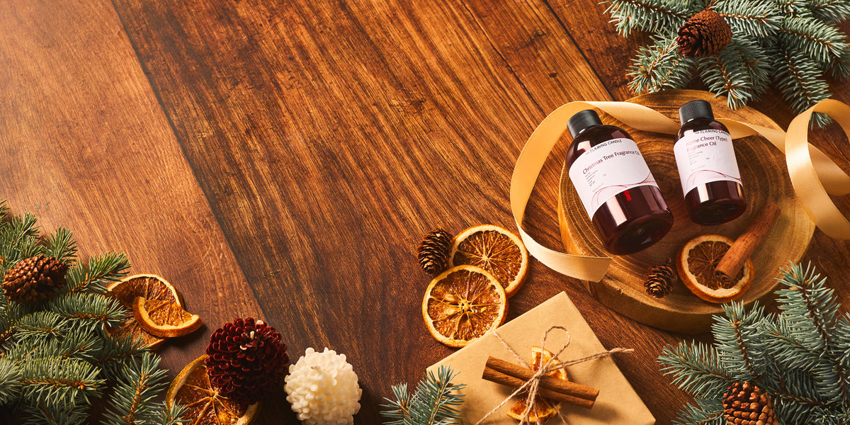 The Flaming Candle Christmas Tree and Alpine Cheer fragrance oils on a wooden tray surrounded by evergreen branches, dried oranges, cinnamon sticks, and pinecones.