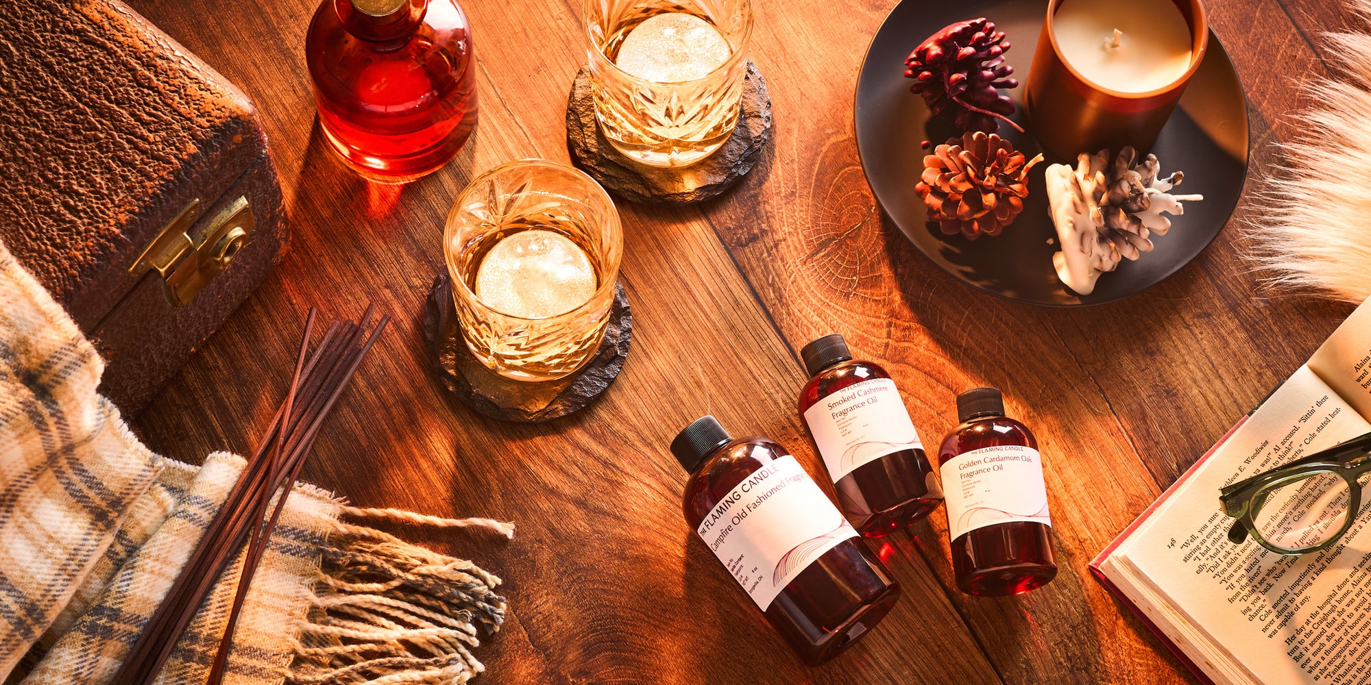 Cozy flat lay with fragrance oil bottles, whiskey glasses on slate coasters, a lit candle, pinecones, reed diffuser sticks, a vintage suitcase, an open book with eyeglasses, and a plaid blanket on a warm wooden surface.