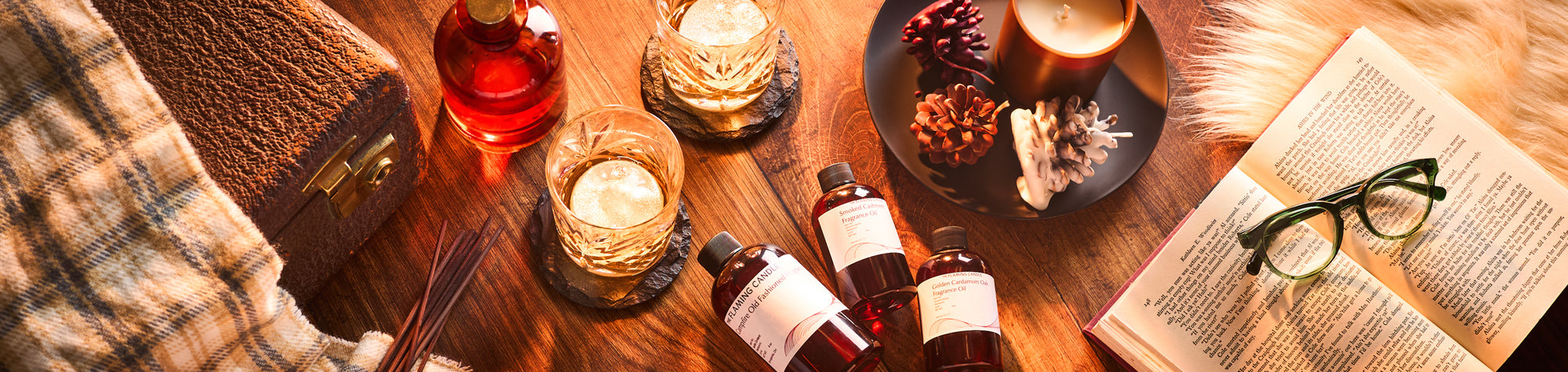 Cozy flat lay with fragrance oil bottles, whiskey glasses, an open book with eyeglasses, a lit candle, pinecones, reed diffuser sticks, a vintage suitcase, and a plaid blanket on a warm wooden surface.