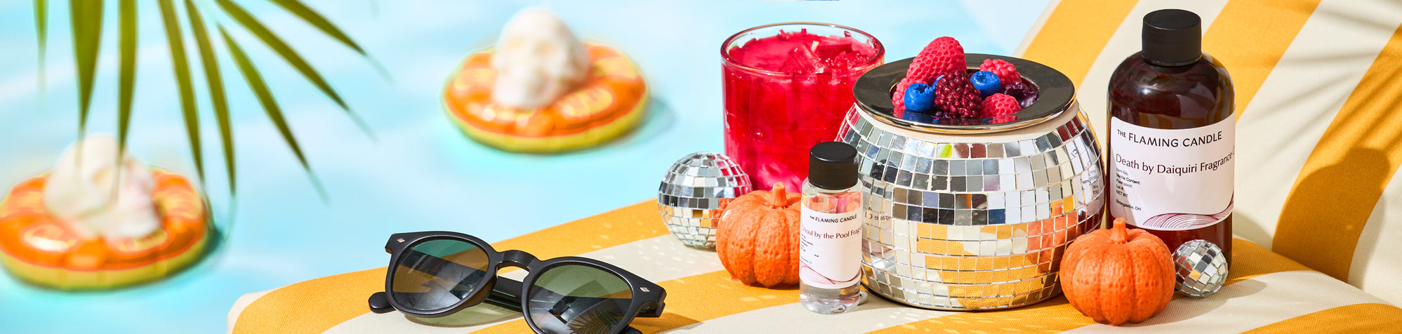 Poolside still life with black sunglasses, a mirrored disco-ball bowl topped with berries, a bright red drink, fragrance bottles, and small pumpkins on a yellow-striped lounge chair beside a blue pool with floating rings.