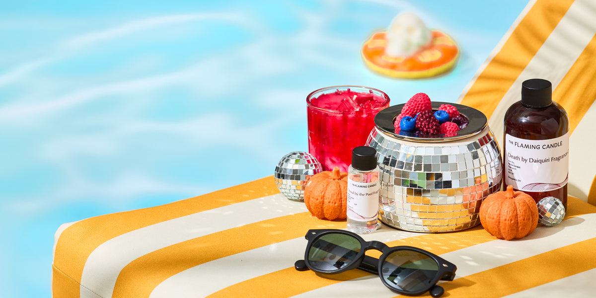 Summerween-themed display by a swimming pool featuring candles, fragrance oils, mini pumpkins, disco ball decor, sunglasses, and berry-topped wax melts on a yellow-and-white striped lounge chair.