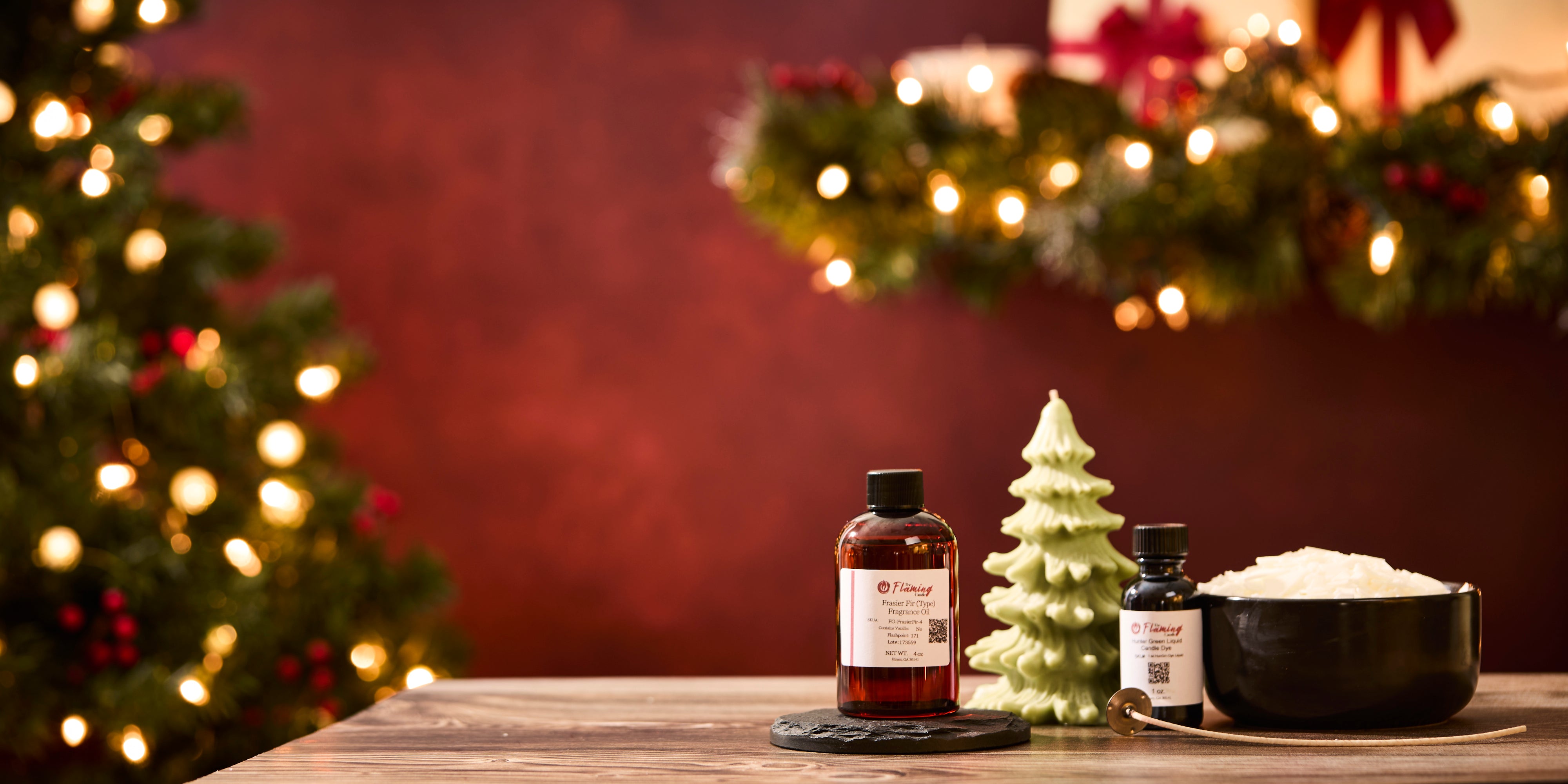 A holiday-themed product scene featuring The Flaming Candle fragrance oils and candle-making supplies arranged on a wooden surface. A red-tinted bottle of fragrance oil sits on a black coaster next to a small green Christmas-tree-shaped candle. A black bowl filled with white wax and a smaller bottle of fragrance oil sit nearby with a wick tool resting in front. In the background, warm Christmas lights, garland, and a decorated tree create a festive, cozy glow.