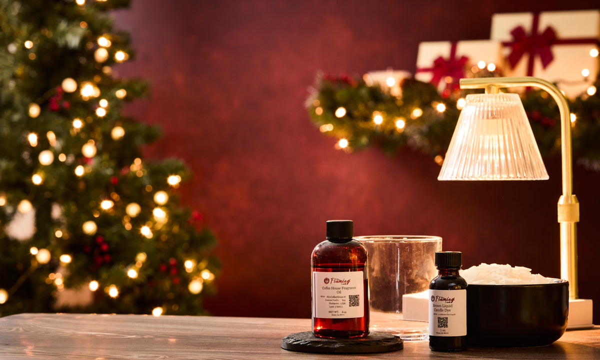 Holiday-themed scene with a blurred Christmas tree and garland lights in the background. On a wooden table in the foreground are candle-making supplies, including a red bottle of The Flaming Candle Coffee House fragrance oil, a small bottle of brown liquid candle dye, a clear empty candle jar, and a black bowl filled with white wax flakes. A gold lamp with a ribbed glass shade glows warmly beside the items.