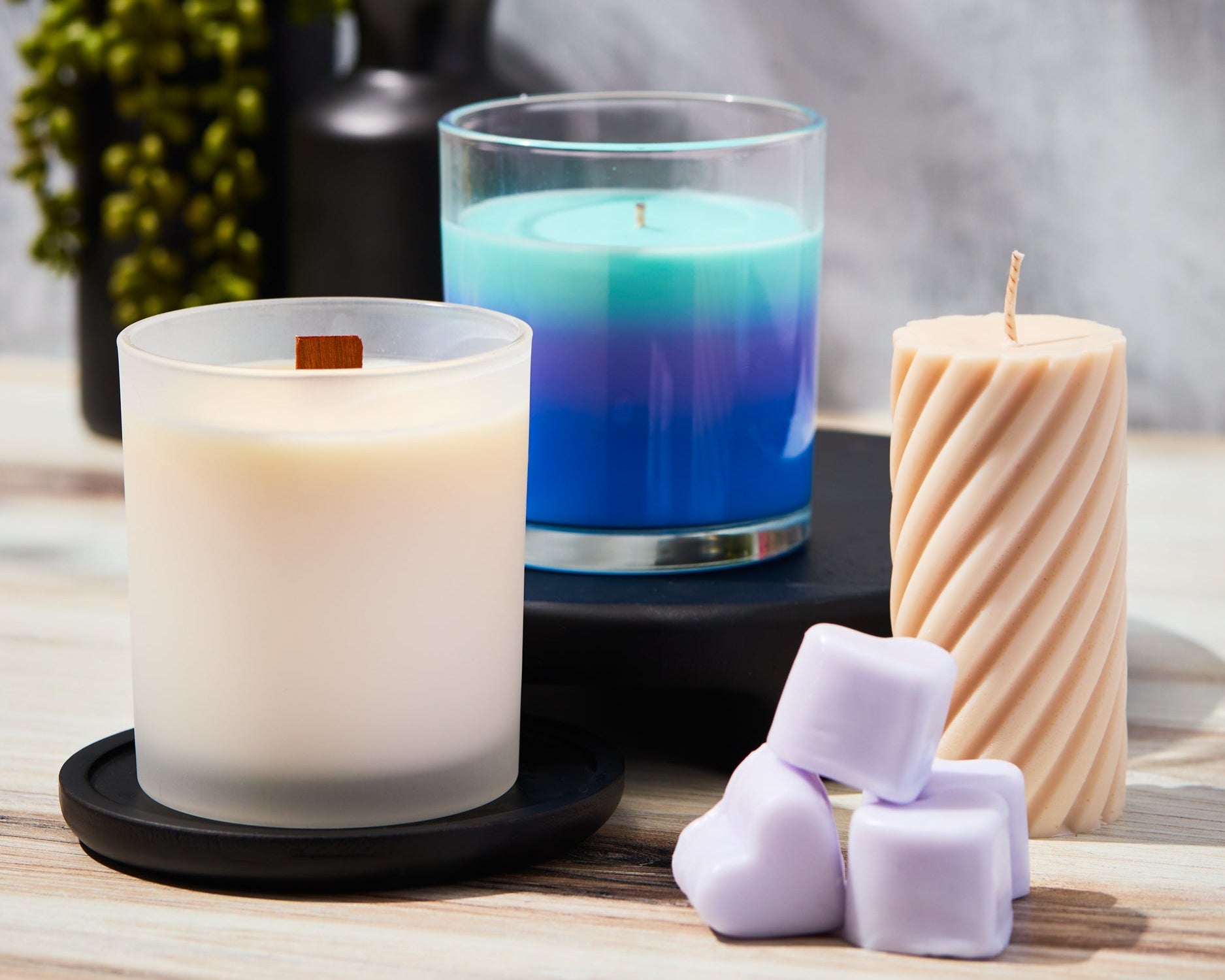 Assorted candles and wax melts displayed on a wooden surface, including a white candle with a wooden wick, a blue layered candle, a twisted pillar candle, and pastel heart-shaped wax melts with decorative greenery and black vases in the background.