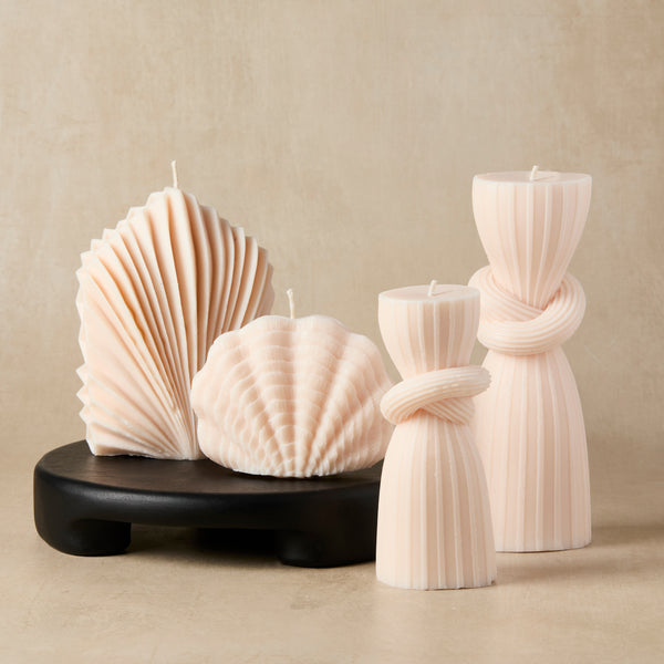 Blush pink sculptural candles with ribbed and shell shapes arranged on a small black pedestal against a neutral beige background.