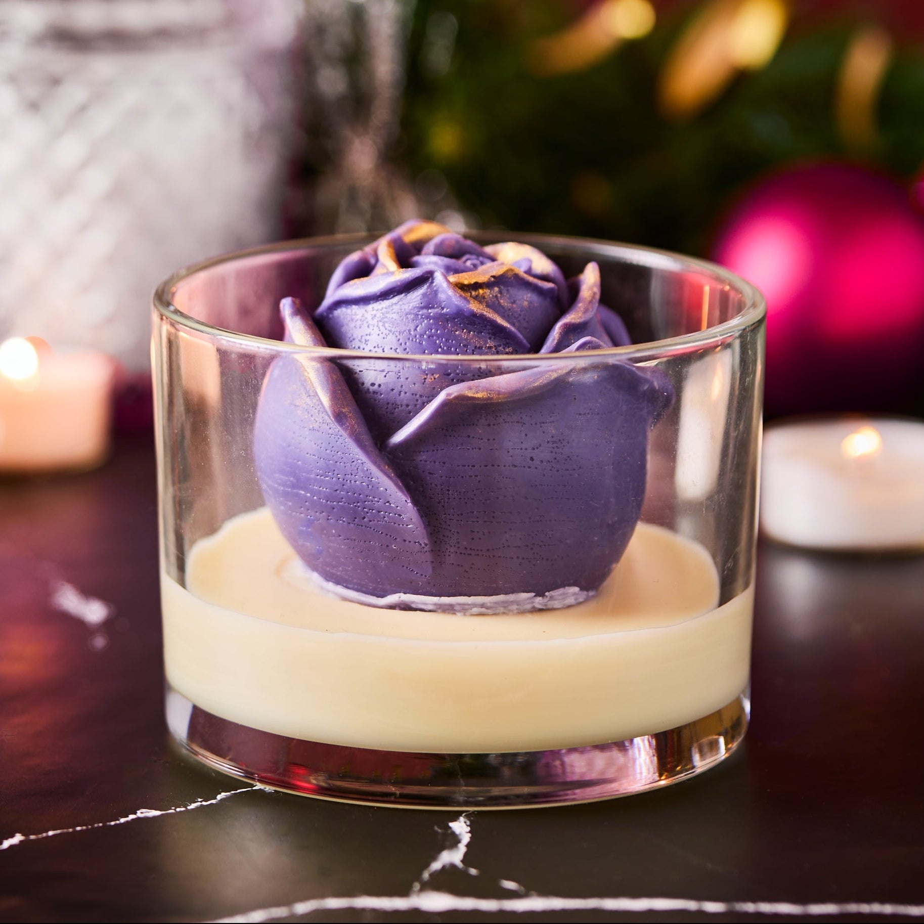 Decorative candle in a glass container featuring a purple rose-shaped wax design, surrounded by tealights and festive ornaments on a dark marble surface.