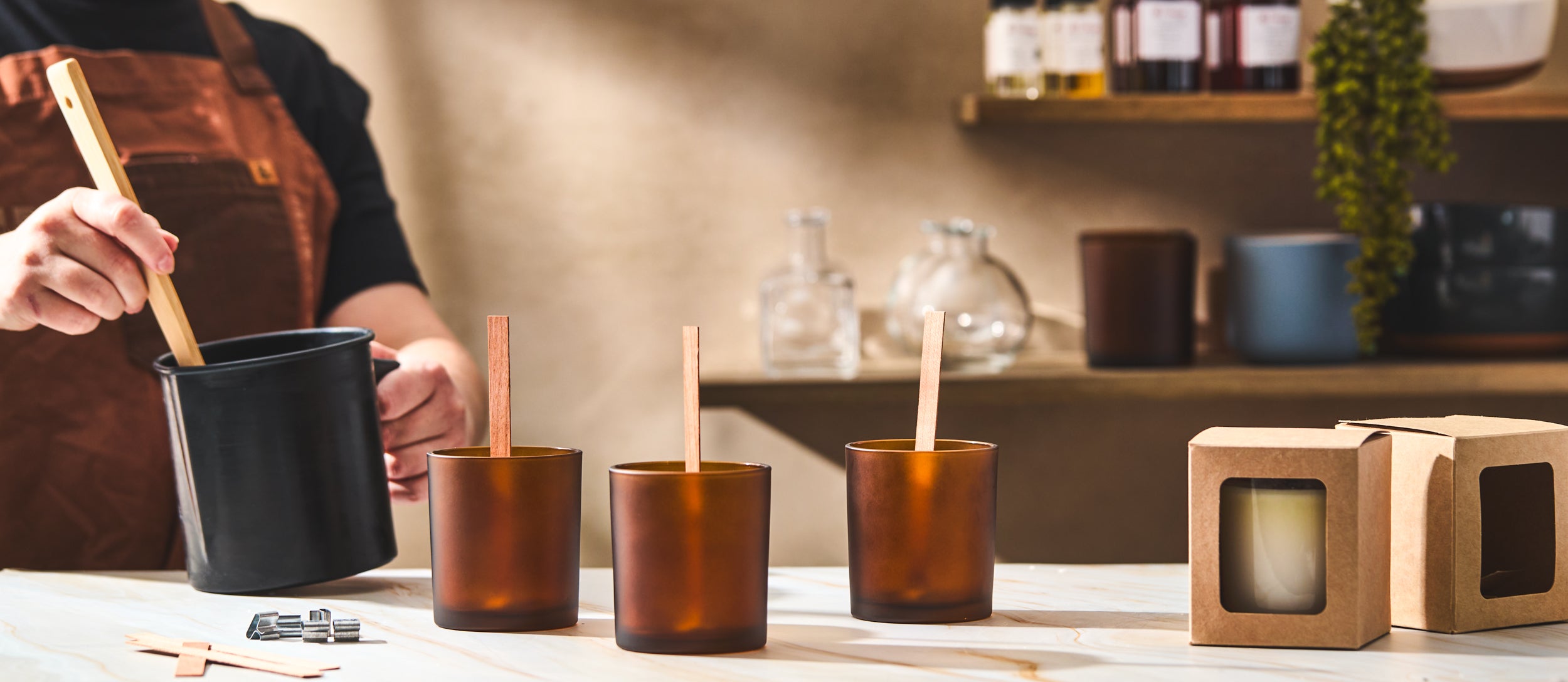 A person wearing a brown apron stirs melted wax in a black pouring pot beside three amber glass candle jars with wooden wicks. Candle packaging boxes and supplies are arranged on the table in a cozy workshop setting.