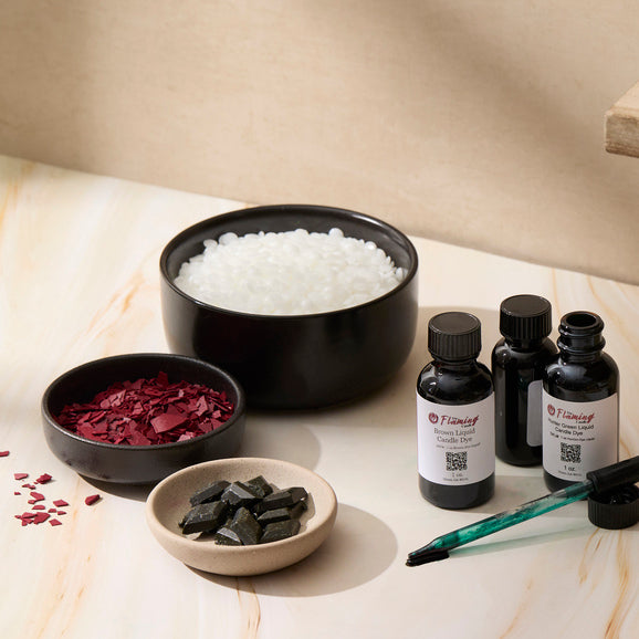 Bowls of wax beads and dye chips are arranged beside bottles of The Flaming Candle liquid candle dye and a dropper, capturing the tools and materials used for custom candle coloring.
