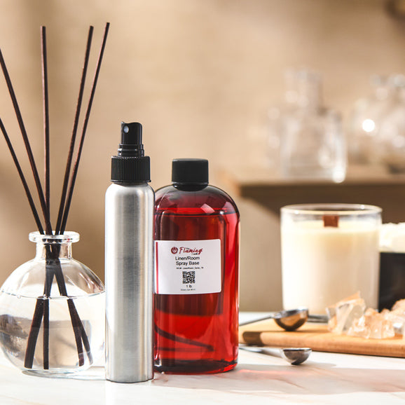 A home fragrance setup featuring The Flaming Candle Linen/Room Spray Base, a silver spray bottle, reed diffuser, and lit candle, styled on a warm, inviting surface with measuring spoons and wax pieces nearby.