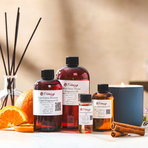 Assorted fragrance oil bottles labeled with The Flaming Candle branding are arranged beside sliced oranges, cinnamon sticks, and a lit candle, creating a warm and aromatic workspace scene.