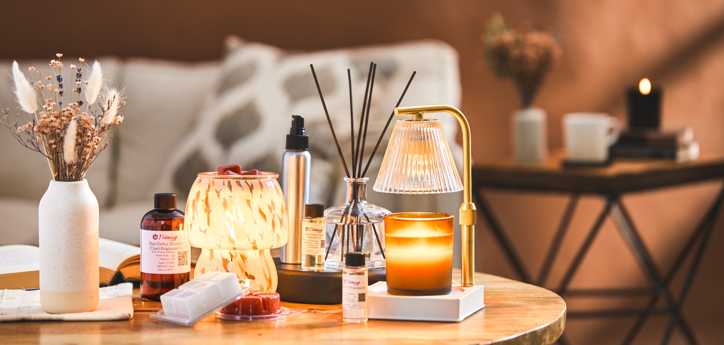 Cozy home scene with The Flaming Candle fragrance oils, wax melts, reed diffuser, and candle warmer on a wooden table beside dried flowers and a glowing candle, creating a warm, inviting atmosphere.